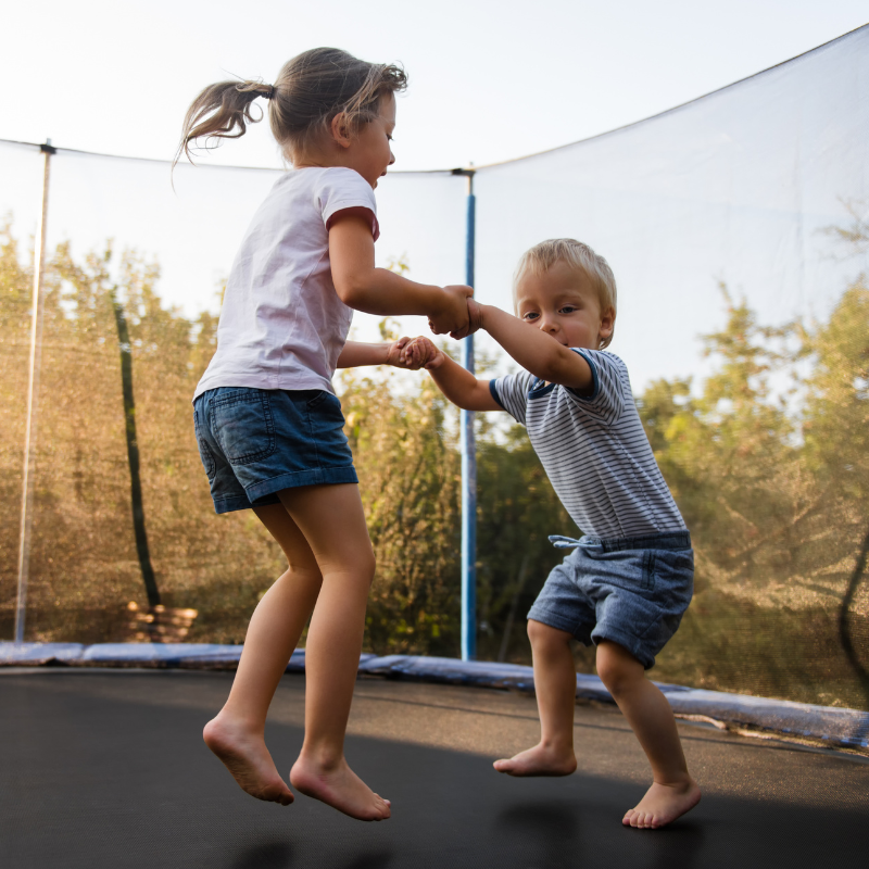 Two young children bounce on a backyard trampoline that has a safety net around it.