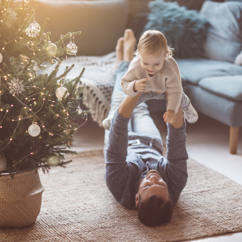 Father on living room floor holding up baby next to holiday tree