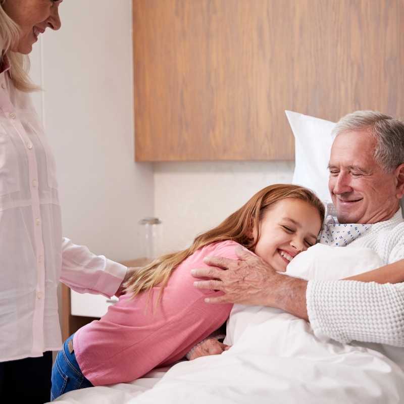older man in hospital bed being hugged by young girl