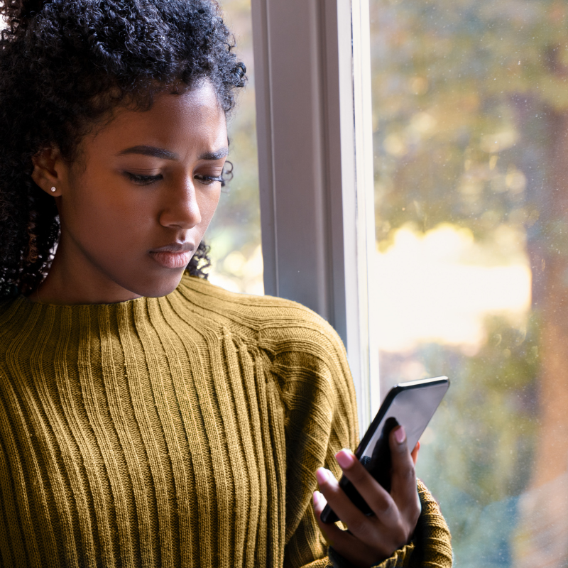 Black woman in yellow sweater with worried expression on her face as she looks at her phone