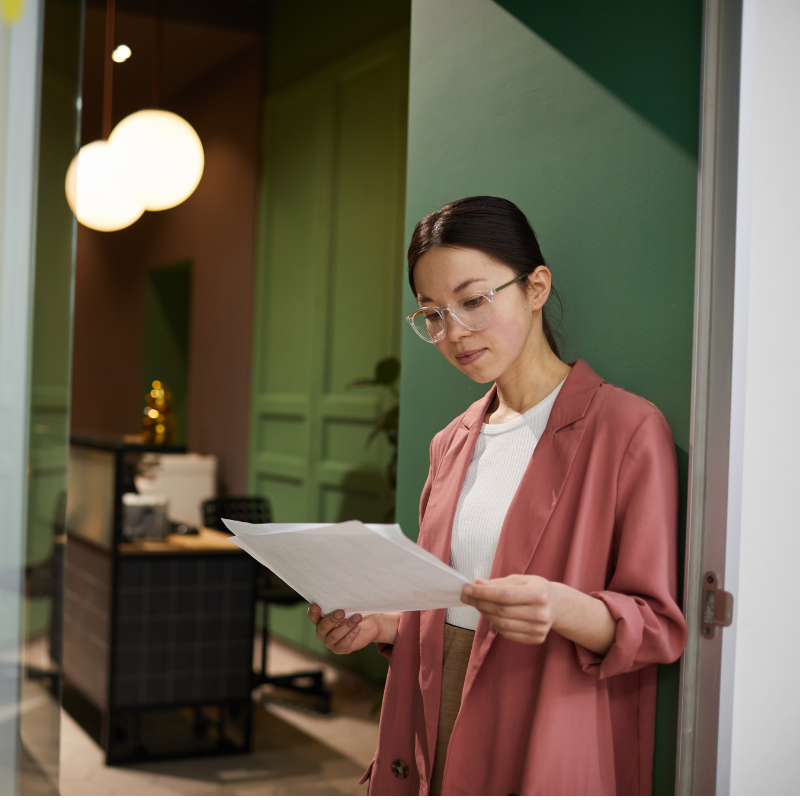 An Asian woman with glasses reads the terms of her car lease. She has her hair pulled into a low ponytail and is leaning against the green wall of her study. 