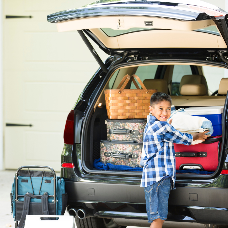 A young tan-skinned boy is putting a bag in a trunk that is already full of luggage, a cooler, and a basket.