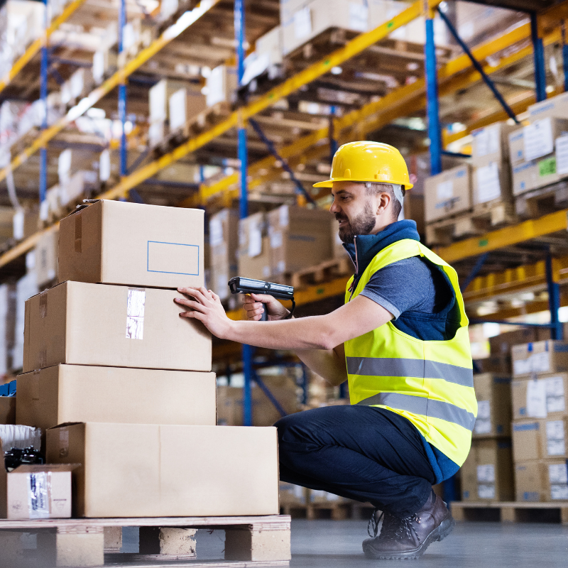 A white male warehouse worker squats down to scan the barcode on a box in a stack. He's wearing a hard had and reflective safety vest, and tall shelves of boxes are behind him.