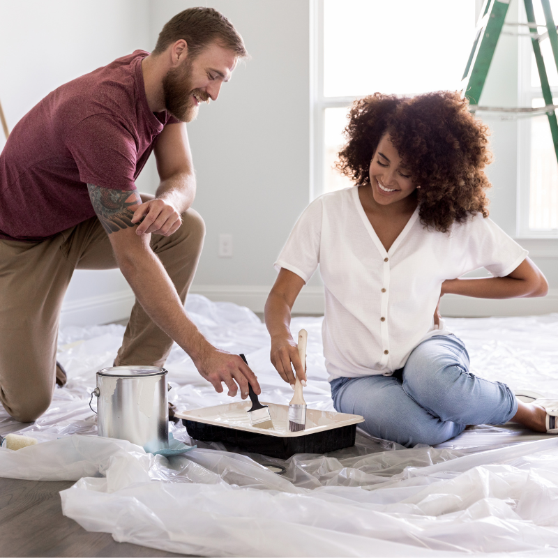 A couple, a white bearded man with tattoos and a curly-haired Black woman, smile as they sit and kneel on the floor dipping brushes in a tray of wall paint. The floor is covered in a tarp and a ladder is set up behind them.
