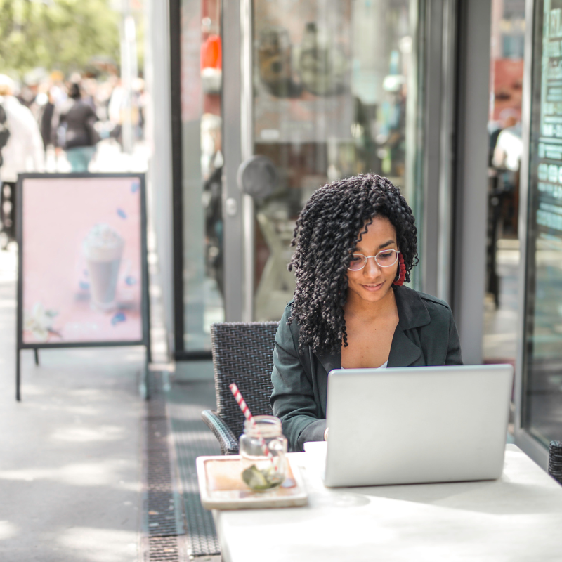 A Black woman with glasses sits outside a city cafe working on her laptop. 