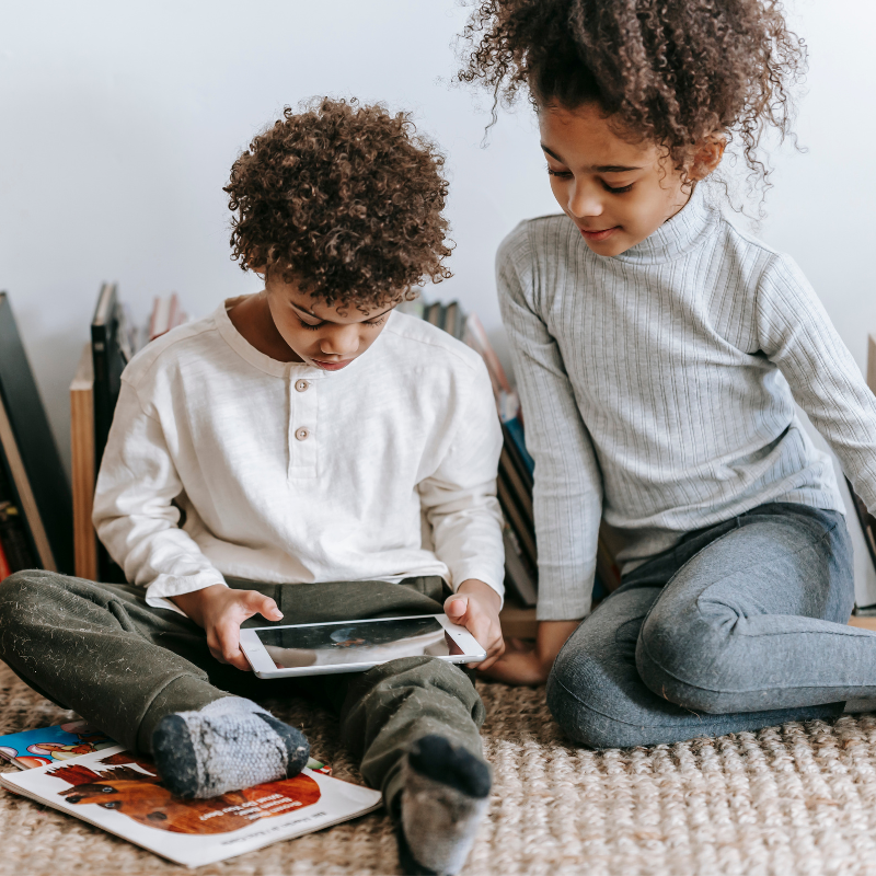 Two young Black children sit on the floor of their playroom and play games together on the family tablet. 