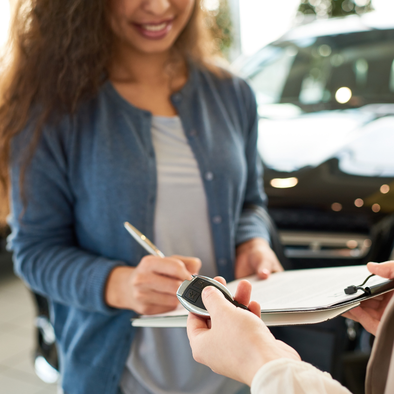 A close-up shot of a curly-haired woman signing for a new car lease. The top half of the woman's face is cropped out, but you can see her smile. Only the salesperson's hands are visible, holding the clipboard and car keys.