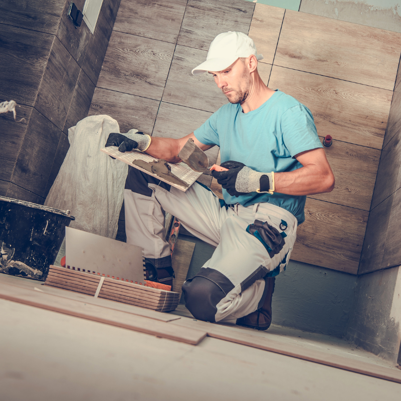 A man crouched on the floor doing tiling work as part of a bathroom remodel.