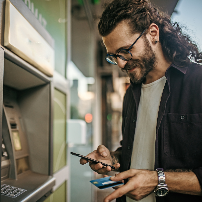 A man with black glasses and his hair pulled back in a ponytail uses an ATM. He's looking at his phone and holding his debit card in his hand.