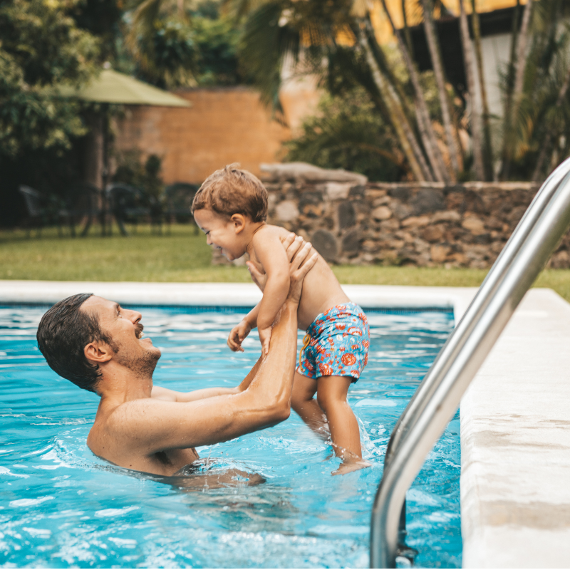 A father plays with his toddler in their backyard in-ground pool. He's holding him up out of the water and they're both smiling and laughing. 