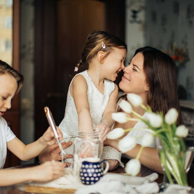 mother rubbing noses with daughter at kitchen table while son stirs a spoon next to them