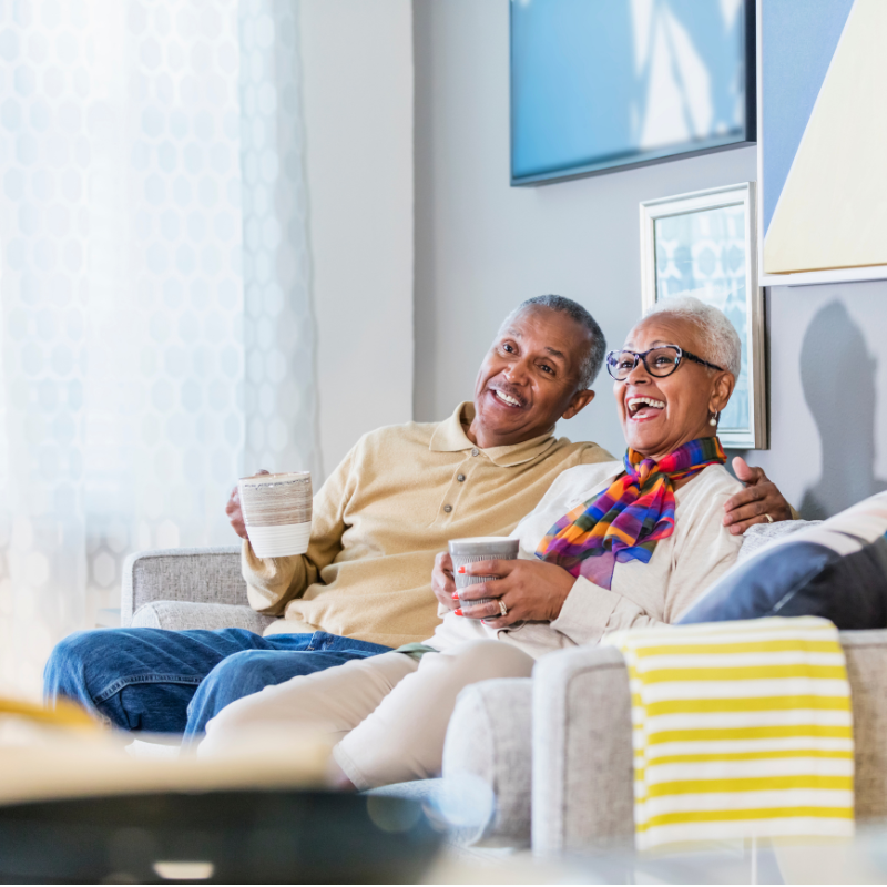 An older Black couple drinking coffee together in their bright living room. 