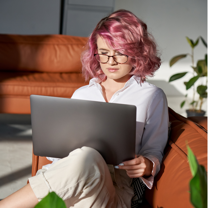 A young woman with pink hair and black glasses works on her laptop in her living room.